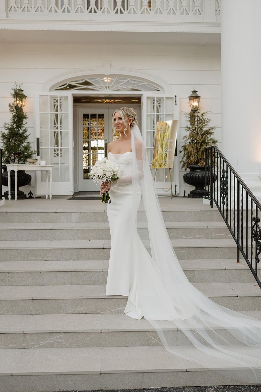 Elegant portrait of the bride posing on steps, holding a white bouquet with a long flowing veil.